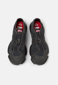 Black athletic shoes with a mesh upper, rubber sole, and padded accents. Features laces and red ‘Camper’ logo on the insoles.
