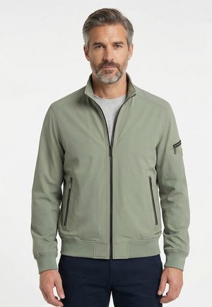 Man met grijs haar en baard, gekleed in een lichtgroene jas met ritssluiting over een grijs shirt en donkere broek, staand tegen een effen achtergrond.