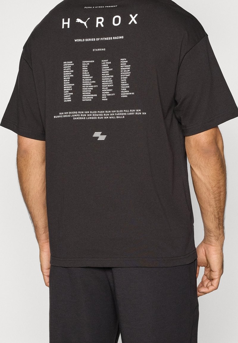 Camiseta de algodón negra con mangas cortas, que presenta un gran texto en blanco en la parte posterior, enumerando ciudades y actividades deportivas relacionadas con HYROX.