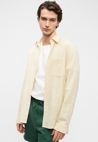 Ελαφρύ κίτρινο πουκάμισο με γιακά και κάθετες ρίγες, μακριά μανίκια, μία τσέπη στο στήθος, φορεμένο πάνω από λευκό T-shirt και πράσινα σορτς.