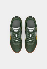 Sapatilhas de camurça verde com detalhes amarelos, atacadores brancos e sola de borracha. Conta com um colarinho almofadado e o logo da Reebok na lingueta.