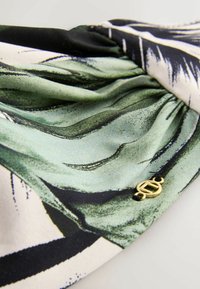 Tessuto verde e nero a motivo con una consistenza setosa, caratterizzato da dettagli arricciati e un piccolo emblema dorato attaccato.