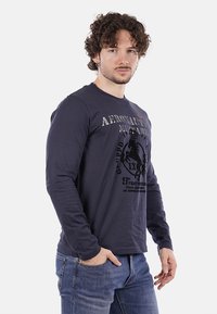 Uomo con capelli ricci che indossa una maglia a maniche lunghe blu navy dell'Aeronautica Militare e jeans blu, in piedi con una mano in tasca.