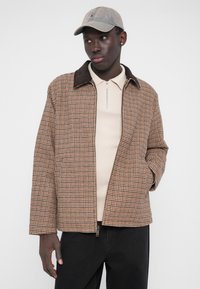 Barna és bézs kockás dzseki barna kordbársony gallérral, elöl cipzárral és két oldalzsebbel, világos bézs felsővel és fekete farmerrel párosítva.