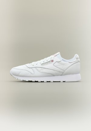 Бели кожени маратонки Reebok с връзки и брандирано лого отстрани и на петата, плоска подметка, на обикновен светлосив фон.