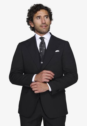 Hombre ajustando el puño de su chaqueta de traje negra, vistiendo camisa blanca y corbata negra con patrón geométrico blanco, mirando ligeramente hacia la derecha.