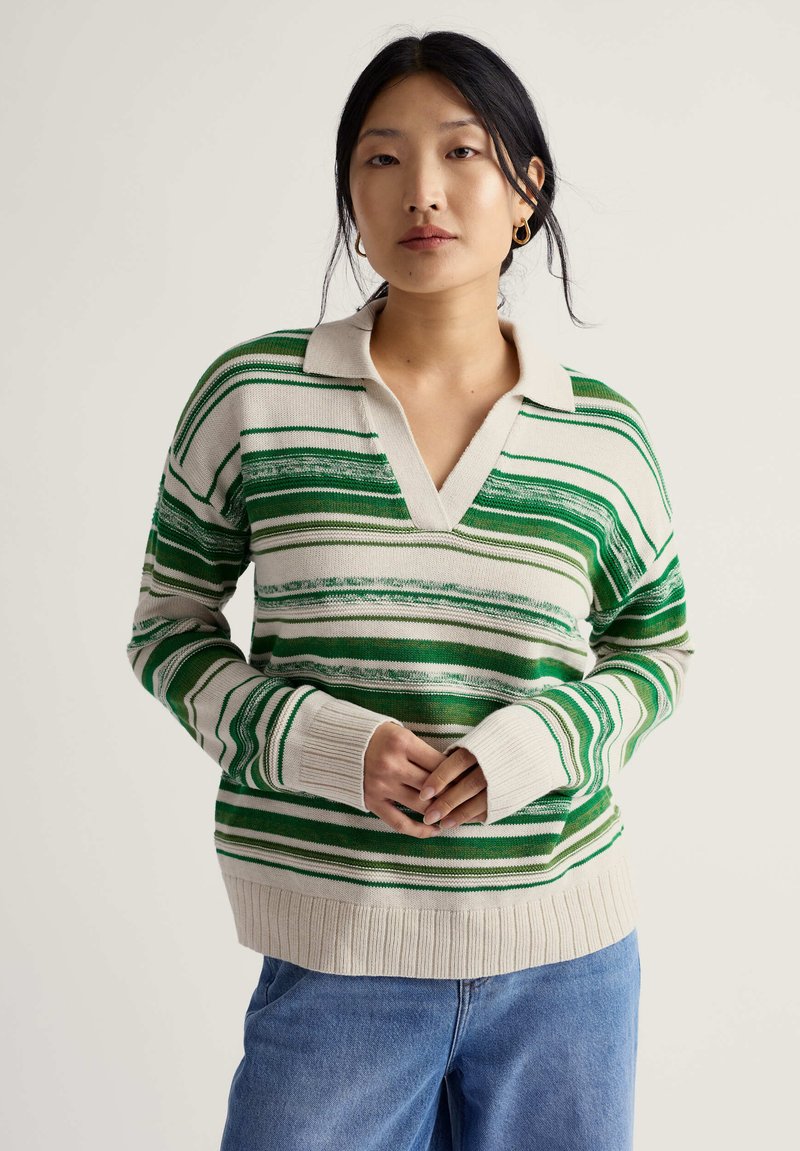 Kvinde med sort hår iført guld øreringe, en creme- og grønstribet sweater med V-hals og blå jeans, stående med sammenknyttede hænder.