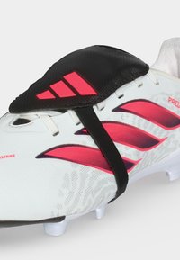 Άσπρο ποδοσφαιρικό παπούτσι με ροζ ρίγες Adidas, μαύρο κάλυμμα κορδονιών με κόκκινο λογότυπο Adidas και λευκές τάπες στη σόλα.