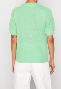 Minzgrüner, gestrickter Kurzarmpullover mit Rundhalsausschnitt, der ein strukturiertes Gewebe und einen gerippten Saum aufweist. Trägt dazu weiße Hosen.