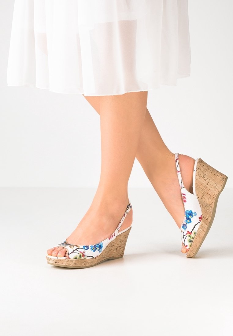 Jambes portant des sandales à talons compensés à bride arrière blanches à motifs floraux avec talons en liège, associées à une jupe blanche fluide sur un fond uni.