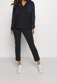 Femme portant une veste matelassée bleu marine, un blouse noire à pois blancs, un pantalon slim noir et des baskets blanches, se tenant contre un fond uni.