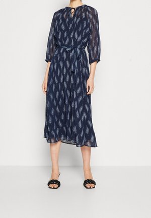 Robe midi bleu marine avec manches longues transparentes et imprimé plumes blanches, taille nouée, encolure en trou de serrure, portée avec des talons ouverts noirs tressés.