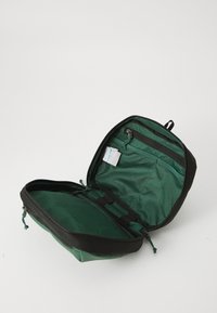 Mochila verde y negra con bolsillos de malla, compartimentos con cremallera y correas ajustables; presenta una forma estructurada y redondeada.