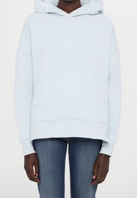 Světle modrá mikina s kapucí, s přední kapsou, oversized střih, žebrované manžety a jemné logo na kapuci. Kombinováno s džíny.