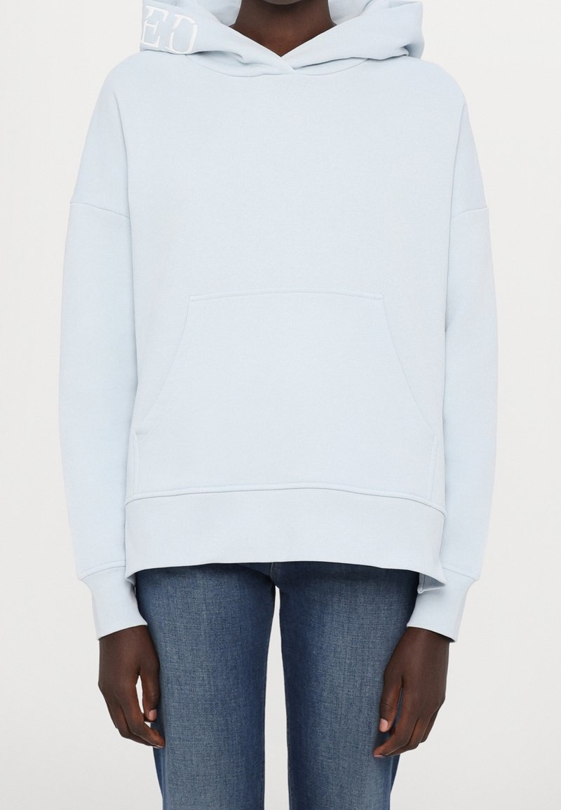 Světle modrá mikina s kapucí, s přední kapsou, oversized střih, žebrované manžety a jemné logo na kapuci. Kombinováno s džíny.