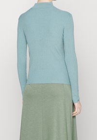 Maglietta a maniche lunghe di un blu chiaro, a costine, con un alto collo, design aderente, che arriva fino alla vita, abbinata a un fondo verde.