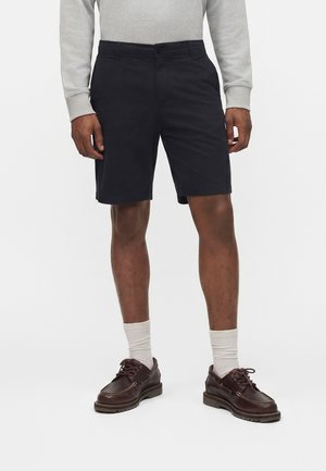 Άνδρας που φοράει μαύρα σορτς μέχρι το γόνατο, γκρι μακρυμάνικη μπλούζα, λευκές κάλτσες και σκούρα καφέ δερμάτινα παπούτσια με κορδόνια, στέκεται μπροστά σε μια απλή φόντο.