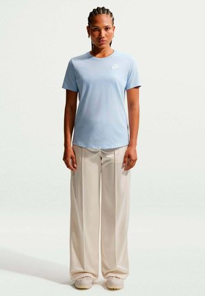 Jonge vrouw met gevlochten haar die een lichtblauw Nike T-shirt, brede beige broek en lichte sneakers draagt, staand tegen een effen witte achtergrond.