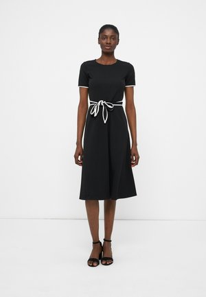 Γυναίκα που φοράει μαύρο φόρεμα μέσης μήκους με λευκή γαζιά στα μανίκια και κορδόνι στη μέση, στέκεται μπροστά σε απλό λευκό φόντο.