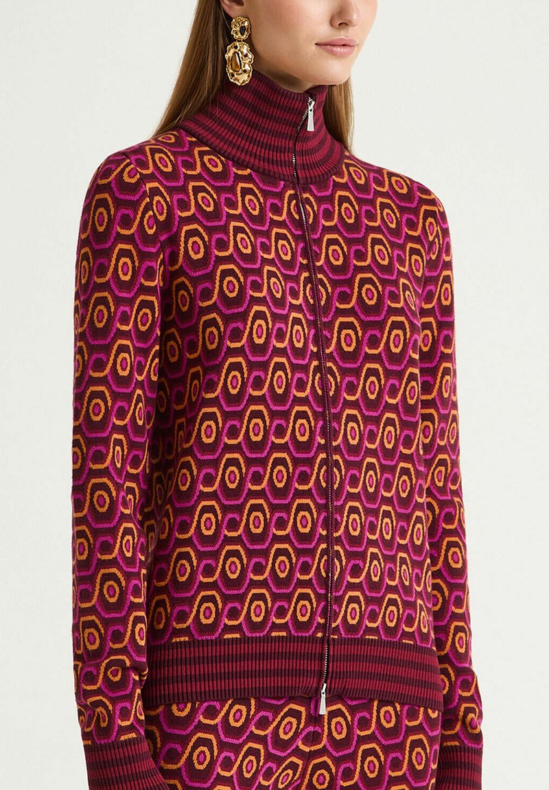 Pull en tricot à fermeture éclair avec un motif géométrique hexagonal en violet, orange vif et bordeaux. Col montant avec des accents côtelés.