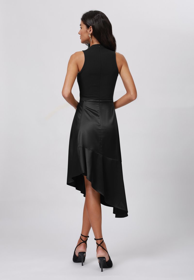 Vrouw met donker haar in een mouwloze zwarte high-low jurk en zwarte gespenhakken, staande tegen een effen lichte achtergrond, achteraanzicht.