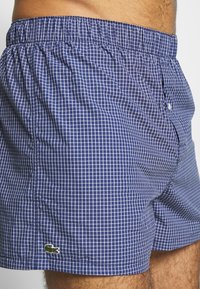 Calções de banho quadriculados em azul-marinho, com cintura elástica e pequeno logótipo da Lacoste. Tecido leve, forma boxy e padrão de grelha branca.