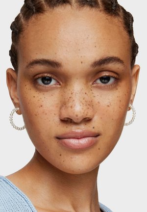 Młoda osoba z zaplecionymi warkoczami, piegami, perłowymi kolczykami kołami i jasnoniebieskim dzianinowym topem, zwrócona przodem na jednolitym tle.