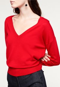 Röd V-ringad tröja med avslappnad passform, ribbad fåll och långa ärmar. Slät yta; bärs med mörk denim för kontrast.