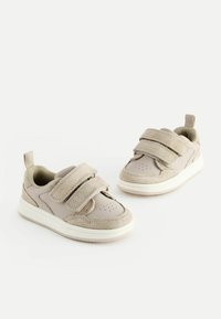 Beige Sneaker, die eine Kombination aus glattem und Wildledermaterial, doppelte Klettverschlüsse, eine perforierte Zehenpartie und eine weiße Gummisohle aufweisen.