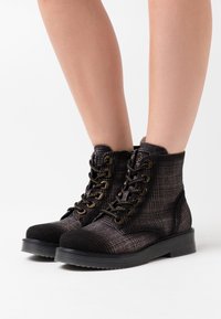 Schwarze, kariert gemusterte Stiefeletten aus strukturiertem Stoff mit runder Zehenpartie, Metallösen und dicker Gummisohle. Die Schnürsenkel verlaufen durch bronzefarbenes Beschlagwerk.