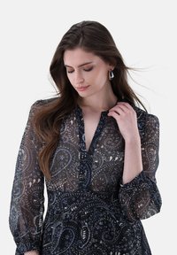 Abito trasparente blu navy con motivo paisley, scollatura a V e maniche lunghe a sbuffo. La modella tiene il colletto, mostrando la leggera consistenza del tessuto.