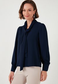 Marineblauwe blouse met een striknek, lange pofmouwen en een gladde, lichte stof. Heeft gathering bij de polsen en een ontspannen pasvorm aan de onderkant.
