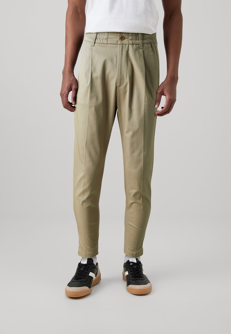 Homme portant un pantalon beige fuselé avec taille élastique et bouton, chemise blanche, baskets noires et beige, debout sur un fond gris uni.