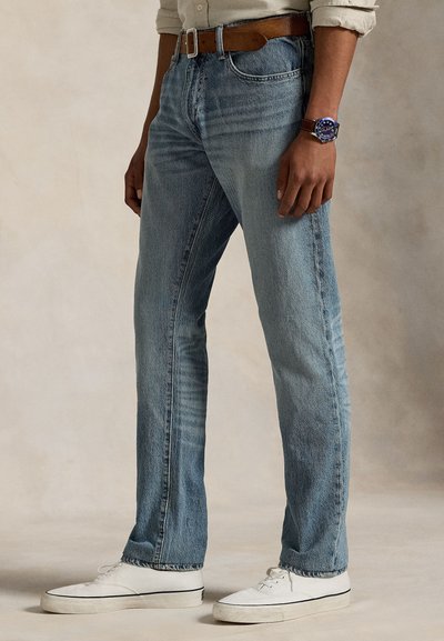 Calças de ganga azul claro com um corte reto, apresentando detalhes desbotados, um cinto de couro castanho e ténis brancos. Vista lateral mostra um corte ajustado.