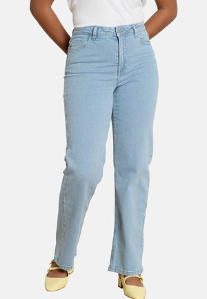 Žena oblečená v svetlomodrých džínsach s rovnými nohavicami s tenkými pásikmi, bielom top a žltých lodičkách na hrubom opätku typu Mary Jane, stojaca na bielom pozadí.