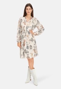 Cremefarbenes Kleid mit Tiermuster, langen Ärmeln und tailliertem Schnitt; kombiniert mit weißen kniehohen, Absatzstiefeln. Glatte Stoffoberfläche.