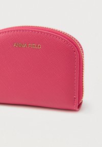 Portefeuille rose zippé avec une surface texturée, des ferrures dorées et un design arrondi. Présente le logo "ANNA FIELD" embossé en or.