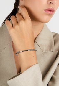 Bracciale in argento su un polso, caratterizzato da un design liscio e arrotondato. Lo sfondo mostra un tessuto beige chiaro con una sottile texture.