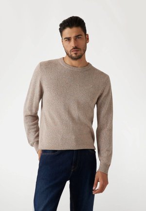 Beige gebreide trui met een ronde halslijn en lange mouwen. De gestructureerde stof heeft subtiele meerkleurige spikkels. Draagt met donkere denim jeans.