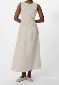 Robe longue sans manches en beige clair avec une taille élastique. Texture lisse, silhouette pleine longueur, assortie à des chaussures slip-on en maille blanche.