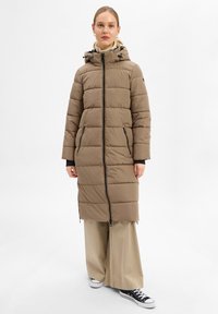 Gevoerde bruine lange jas met een capuchon, voorrits, twee zijzakken en geribde manchetten, gecombineerd met beige wijde broek en zwarte sneakers.