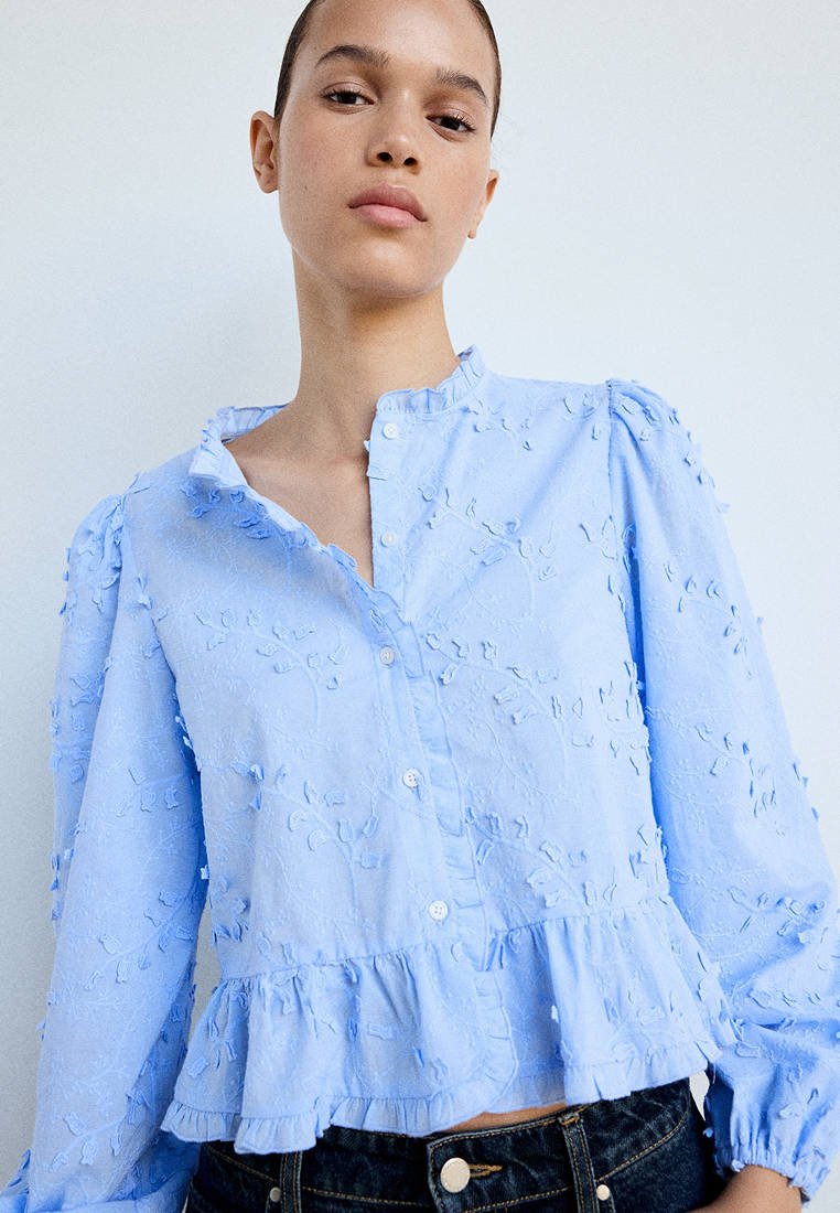Jeune personne portant un chemisier bleu clair texturé avec un ourlet volanté et des manches longues, associé à un jean denim foncé, sur un fond uni.