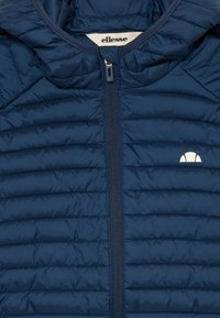 Veste matelassée bleu marine avec fermeture éclair à l'avant et logo ellesse blanc sur la poitrine, présentant un motif de couture horizontale rembourrée.