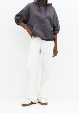 Personne portant un sweat à capuche oversize gris foncé, un pantalon blanc large texturé et des baskets blanches avec des semelles marron, debout devant un fond blanc.