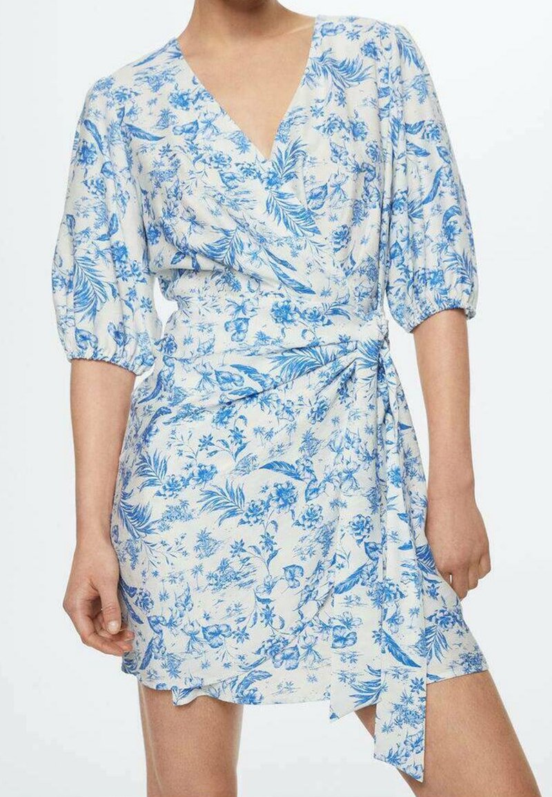 Robe portefeuille blanche avec un motif floral et de feuilles bleues, col en V, manches bouffantes mi-longues et ceinture nouée à la taille sur une personne debout.