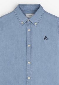 Camisa de mezclilla azul claro con botones, cuello plano, botones blancos y un pequeño logo de calavera y tibias cruzadas en el lado izquierdo del pecho.