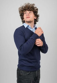 Námořnická modrá pletená svetr s výstřihem do V, oblečený přes světle modrou košili s límcem, na osobě, která upravuje svetr.