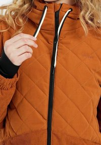 Gepolsterte braune Jacke mit hohem Kragen, ausgestattet mit einem schwarzen Reißverschluss, weißen Zugbändern und gerippten Bündchen für zusätzlichen Komfort.