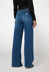 Plačios kojų mėlynos džinsinės kelnės su aukštu liemeniu, su dviem užpakalinėmis kišenėmis ir maža trikampio formos logotipų etikete virš dešinės kišenės.