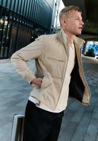 Lichtbeige ritsjack met een hoge kraag, voorzak en elastische manchetten, gedragen over een wit shirt en zwarte broek. Glad textuur.